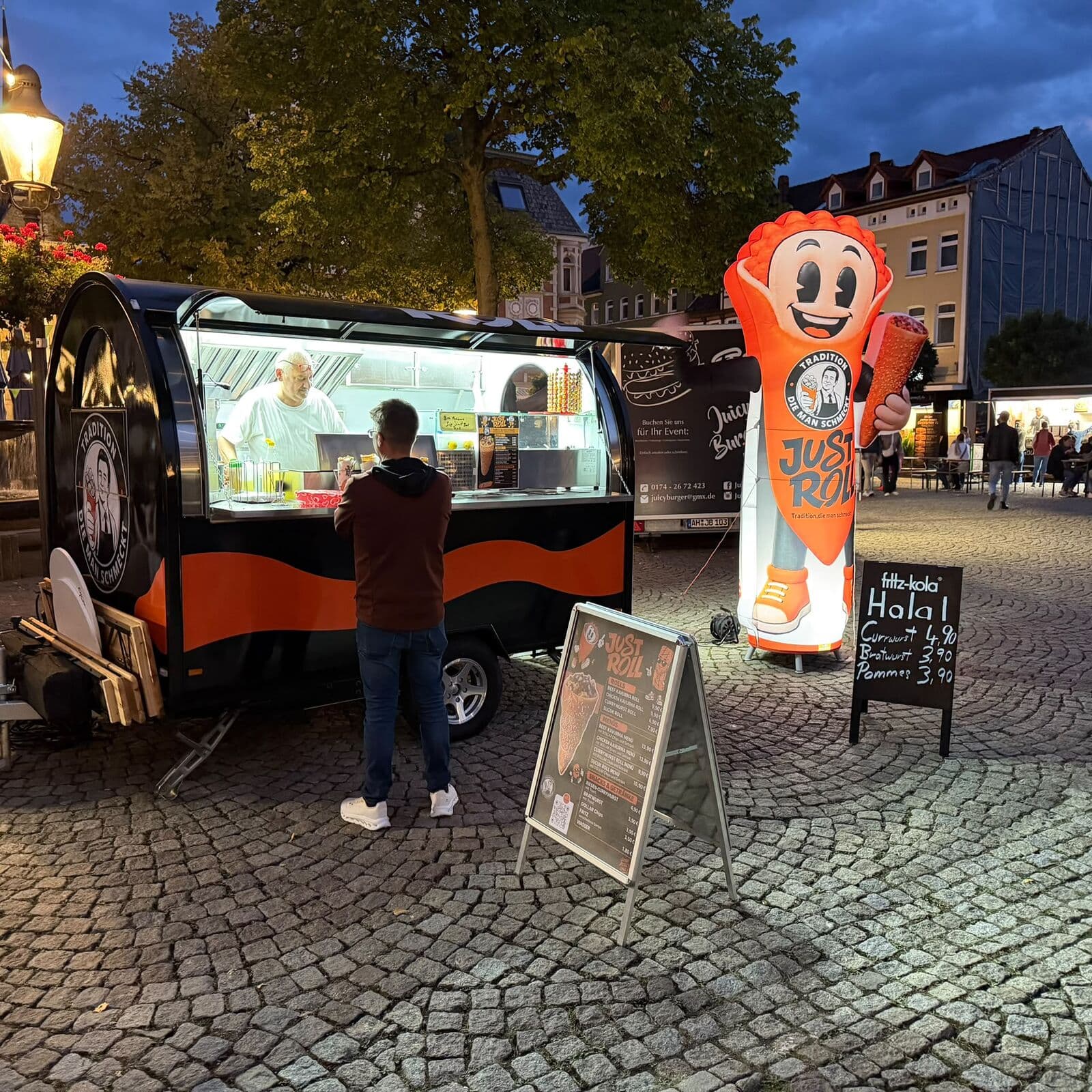 Just-Roll-Trailer am Abend auf einem Marktplatz, ein Gast steht an der Ausgabe